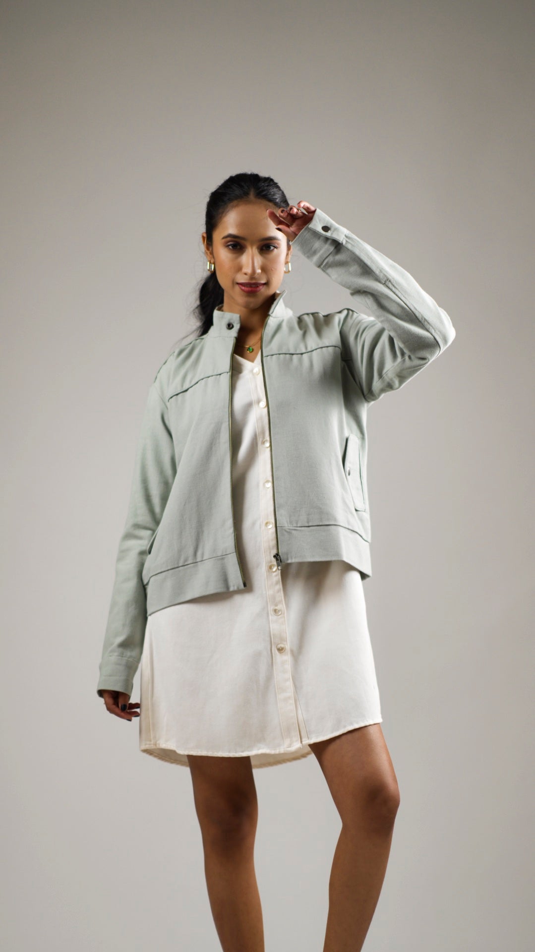 A woman is posing with one hand raised to her head. She is wearing a long, olive green cotton canvas bomber jacket with a stand collar, functional welt pockets with flaps, and snap buttons on the collar and pockets.