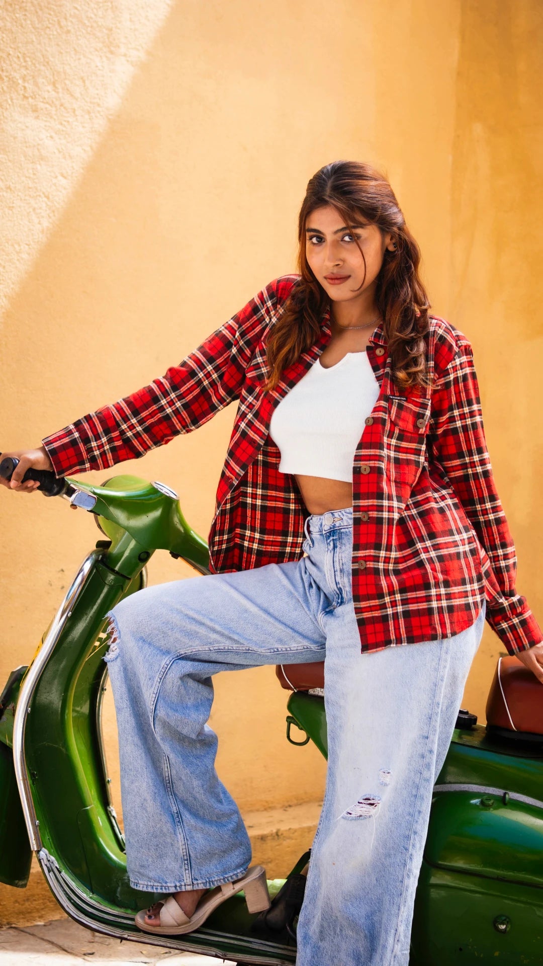 Woman wearing a red plaid shirt, white top, and blue jeans sitting on a green scooter against a beige wall.