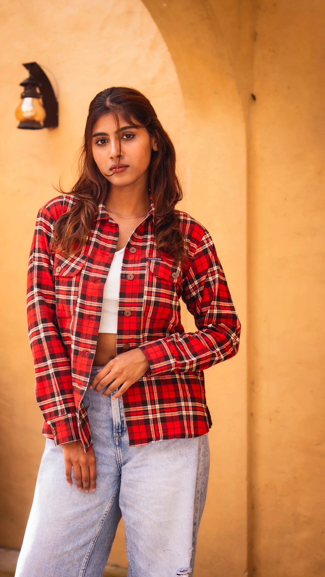Woman wearing a red plaid shirt and light blue jeans against a beige wall.