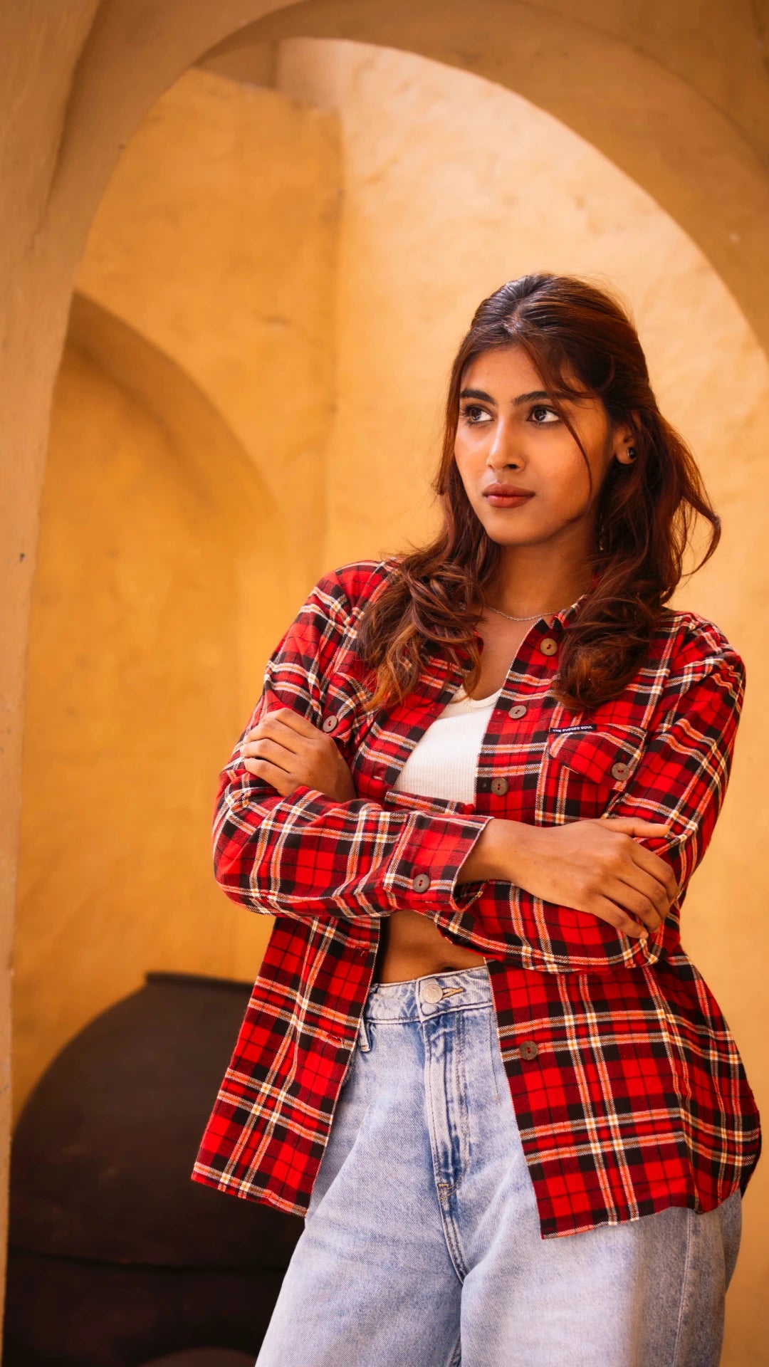 Woman wearing a red plaid shirt over a white top and blue jeans, standing against a beige wall.