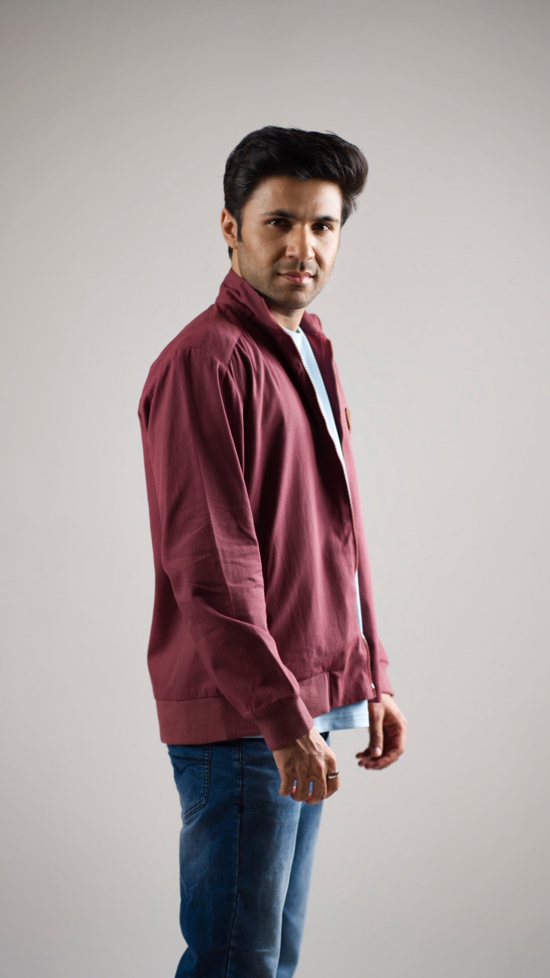 A man wearing a deep garnet-coloured cotton twill jacket with a stand collar, discreet welt pockets, and ribbed cuffs. He is also wearing a light shirt and blue jeans.