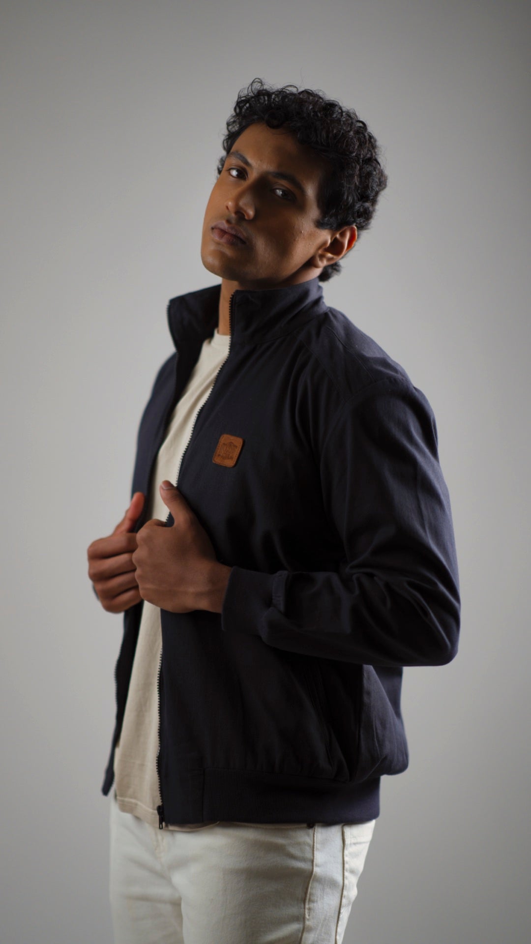 A man wearing a midnight navy cotton twill jacket with a stand collar, discreet welt pockets, and ribbed cuffs. He is paired with a cream shirt and light-coloured trousers.