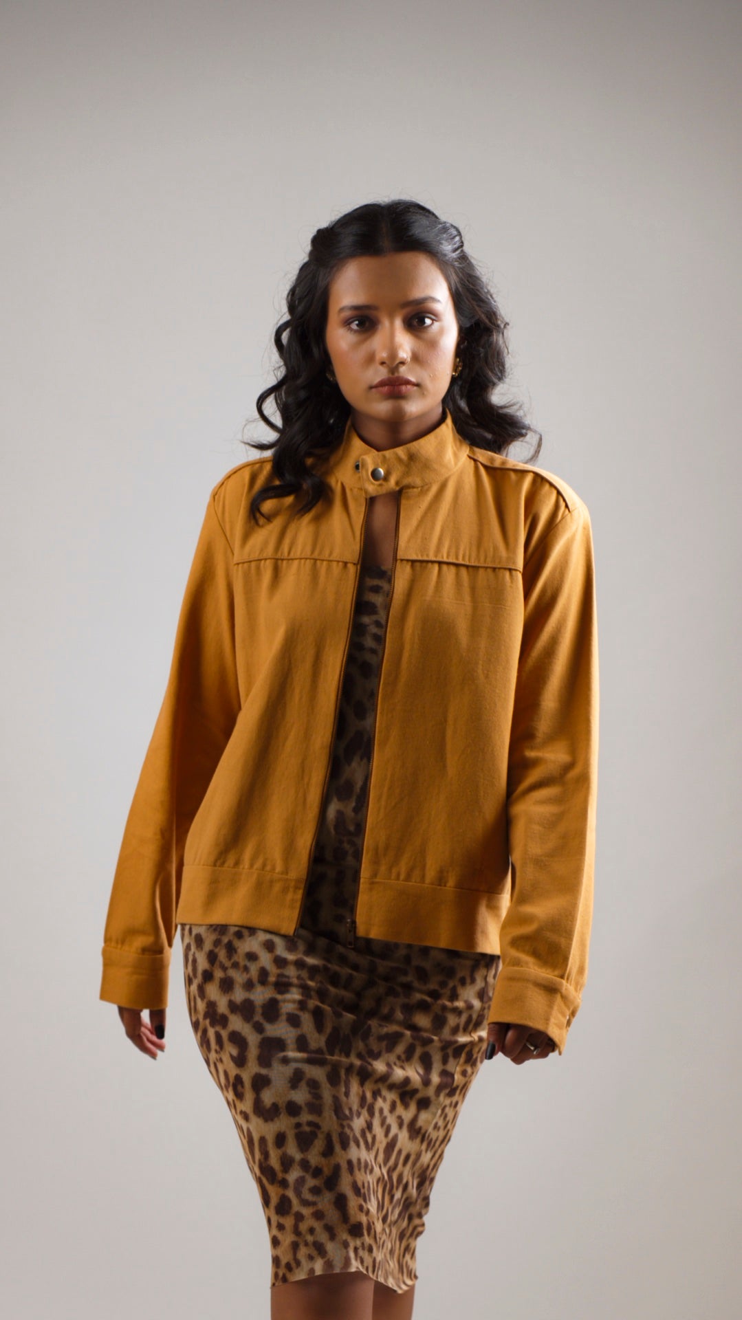 A woman standing in a studio setting, wearing a caramel tan bomber jacket with a stand collar, functional welt pockets with flaps, and snap buttons. She is also wearing a leopard print skirt.