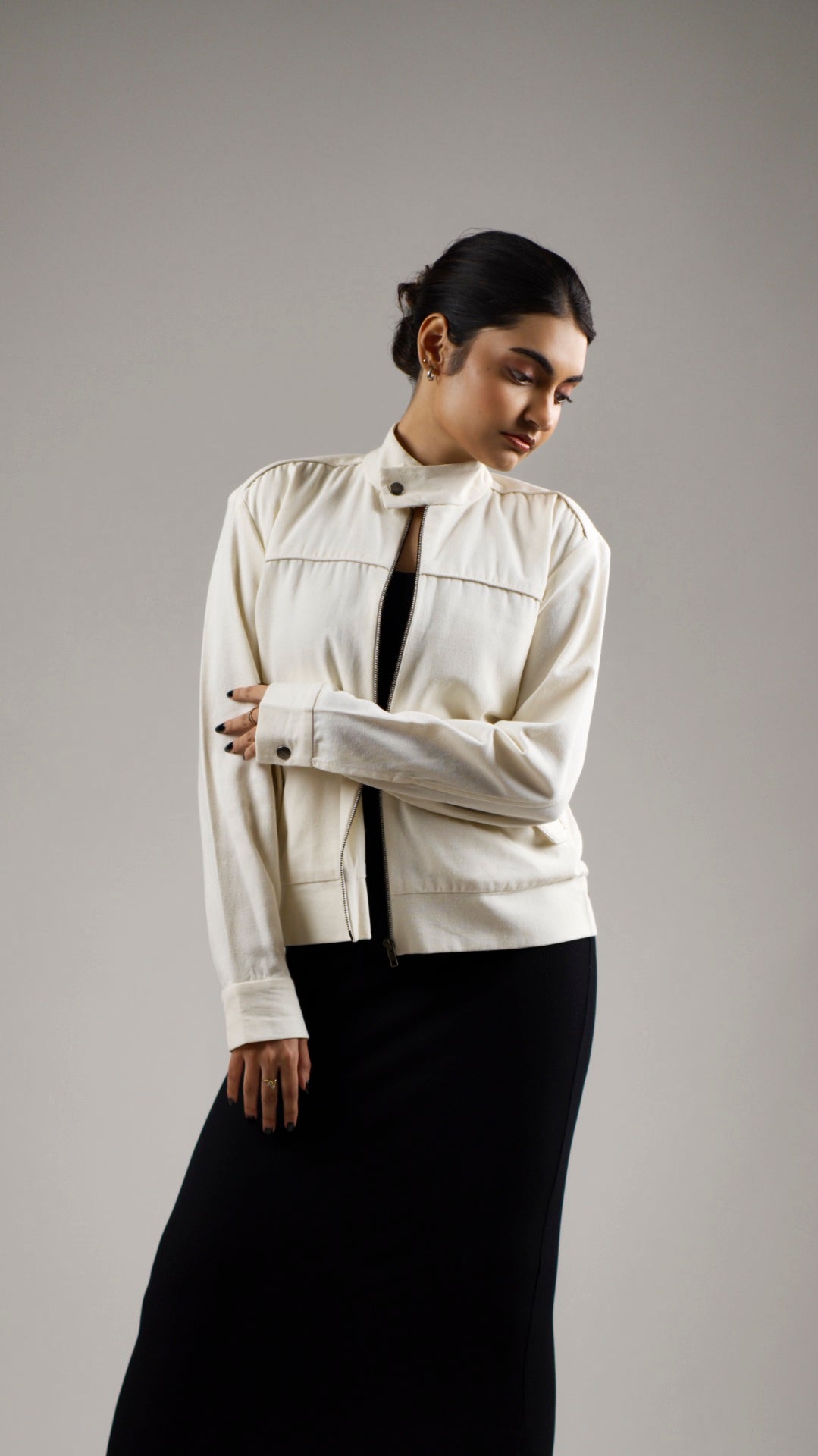 A woman wearing a vanilla cream colored cotton canvas jacket with a stand collar, functional welt pockets with flaps, and snap buttons on the collar and pockets. She is also wearing a dark skirt.