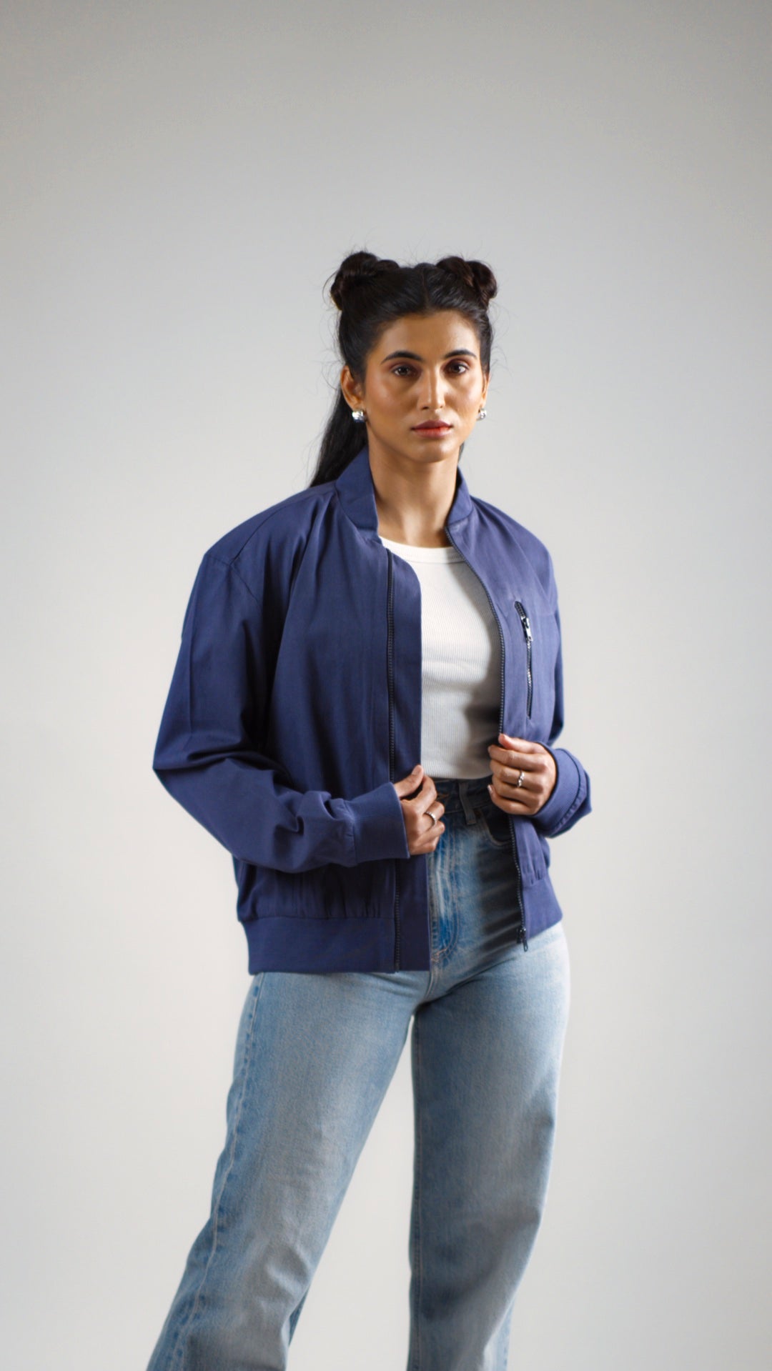 A woman standing against a plain background, wearing a steel blue cotton twill jacket with round neckline, sleeve cuffs, and diagonal front pockets. She is also wearing light blue jeans.