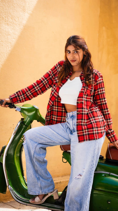 Woman wearing a red plaid shirt, white top, and blue jeans sitting on a green scooter against a beige wall.